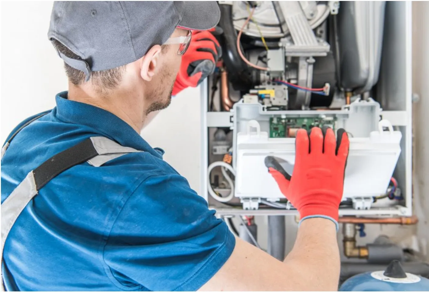A technician in a blue shirt and red gloves works on a boiler, adjusting components inside the open panel.
