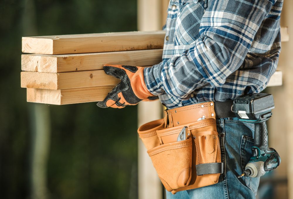 A person in plaid shirt carries wooden planks, wearing gloves and a tool belt with a drill on the side.