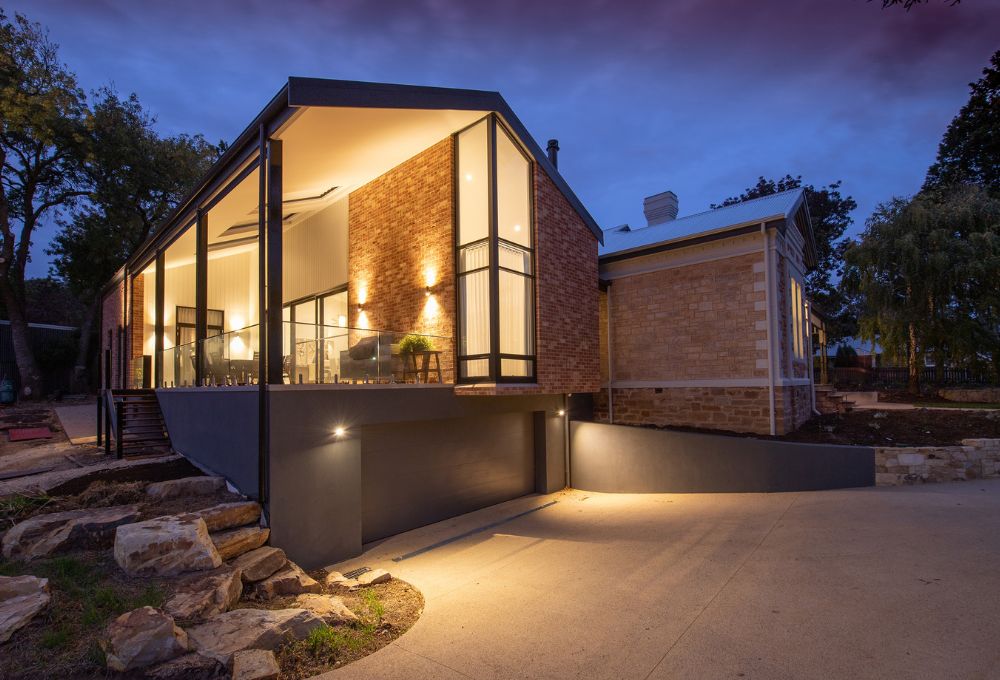 Modern house with large windows and warm lighting, surrounded by trees, at dusk.