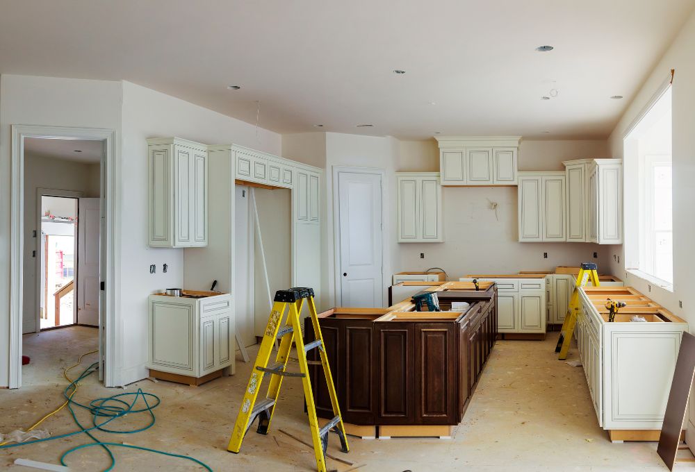 Unfinished kitchen with white cabinets, brown island, ladders, and tools on a dusty floor, under bright natural light.