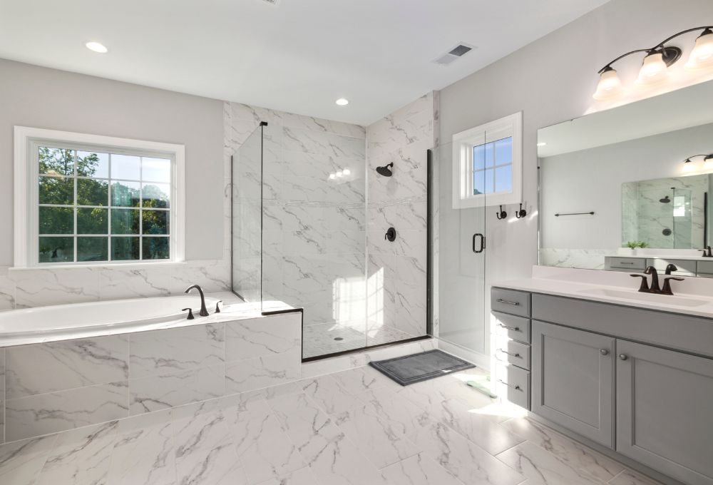 A modern bathroom with marble tiles, a glass shower, a bathtub, a large mirror, and gray cabinets under bright lighting.