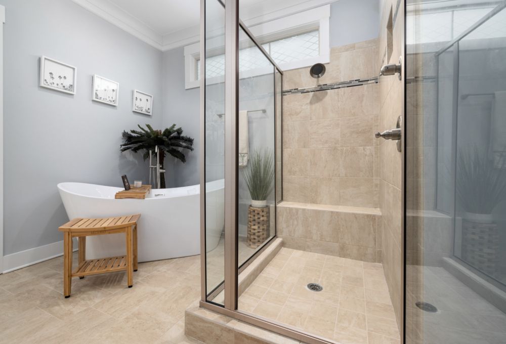 Modern bathroom with a glass shower, freestanding tub, wooden stool, and decorative plants on beige tiled floor.