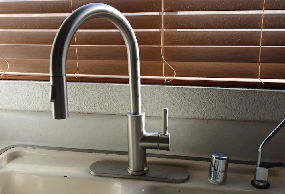 A stainless steel kitchen faucet with a curved spout is set against closed wooden blinds.