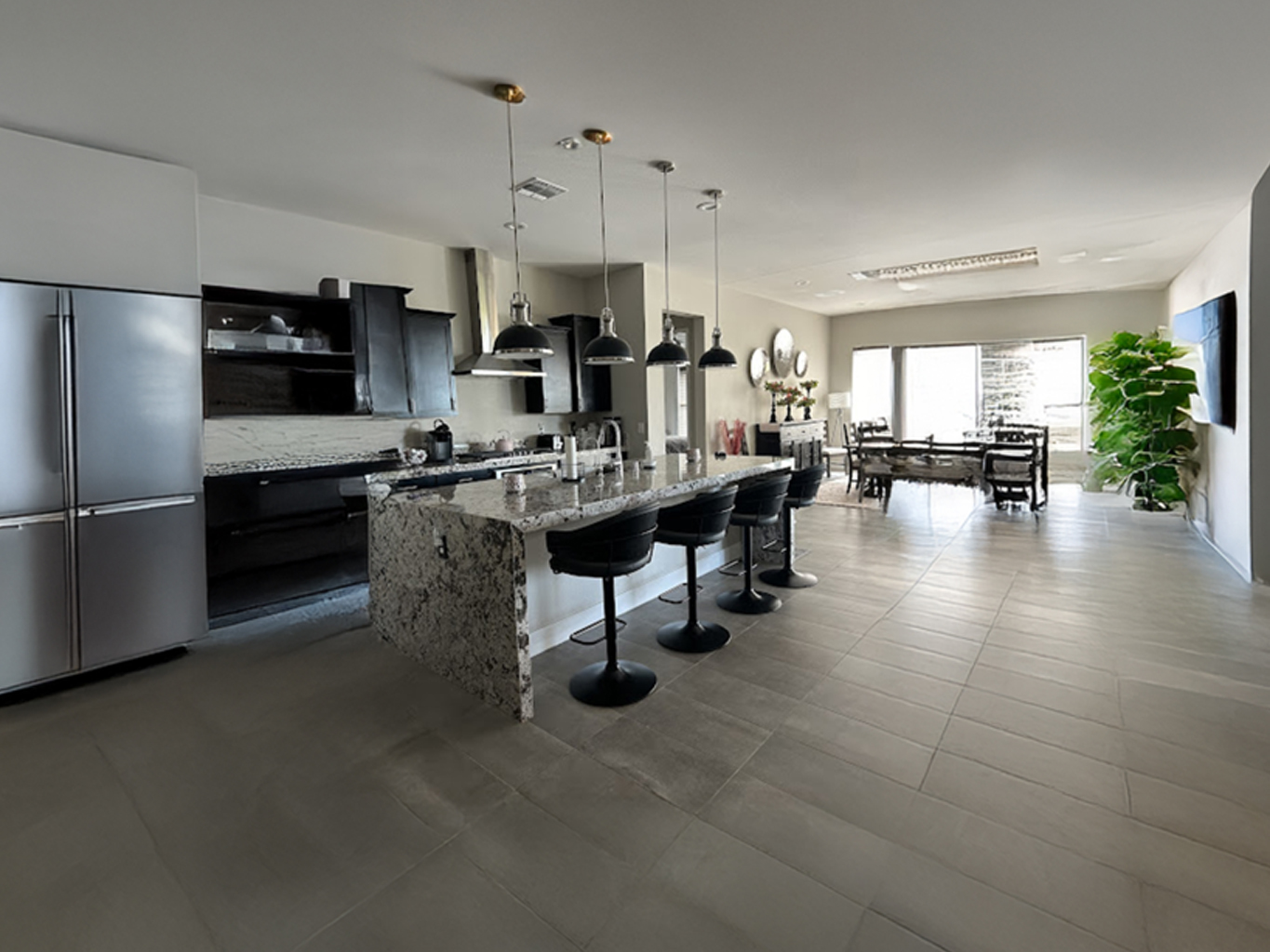 Modern kitchen with granite island, pendant lights, bar stools, dining table, and large plant near bright windows.